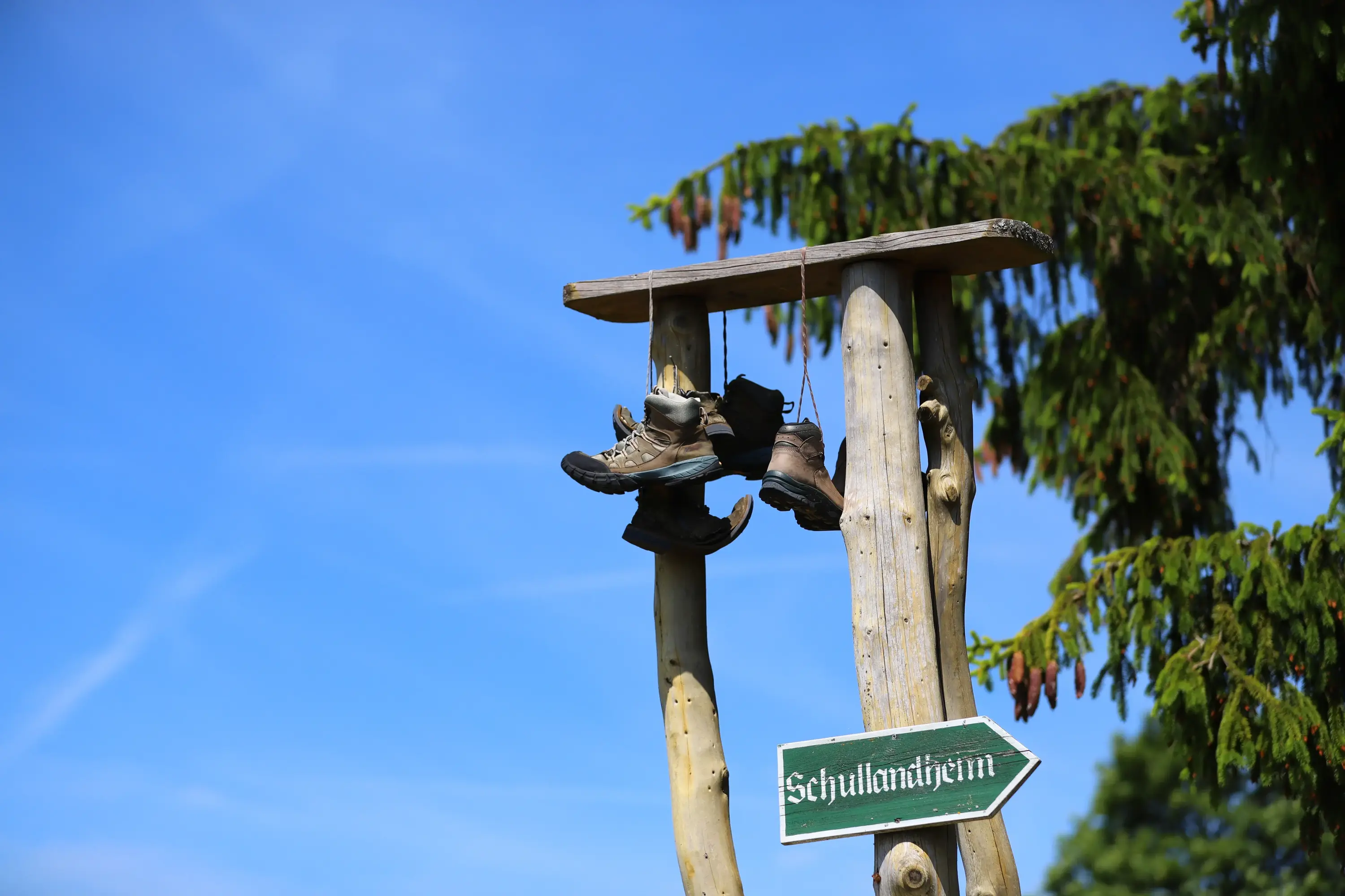 Schuhe an den Haken Ausgediente Wanderschuhe hängen an einem Holzgestell vor blauem Himmel, daneben ein grüner Wegweiser mit der Aufschrift Schullandheim.