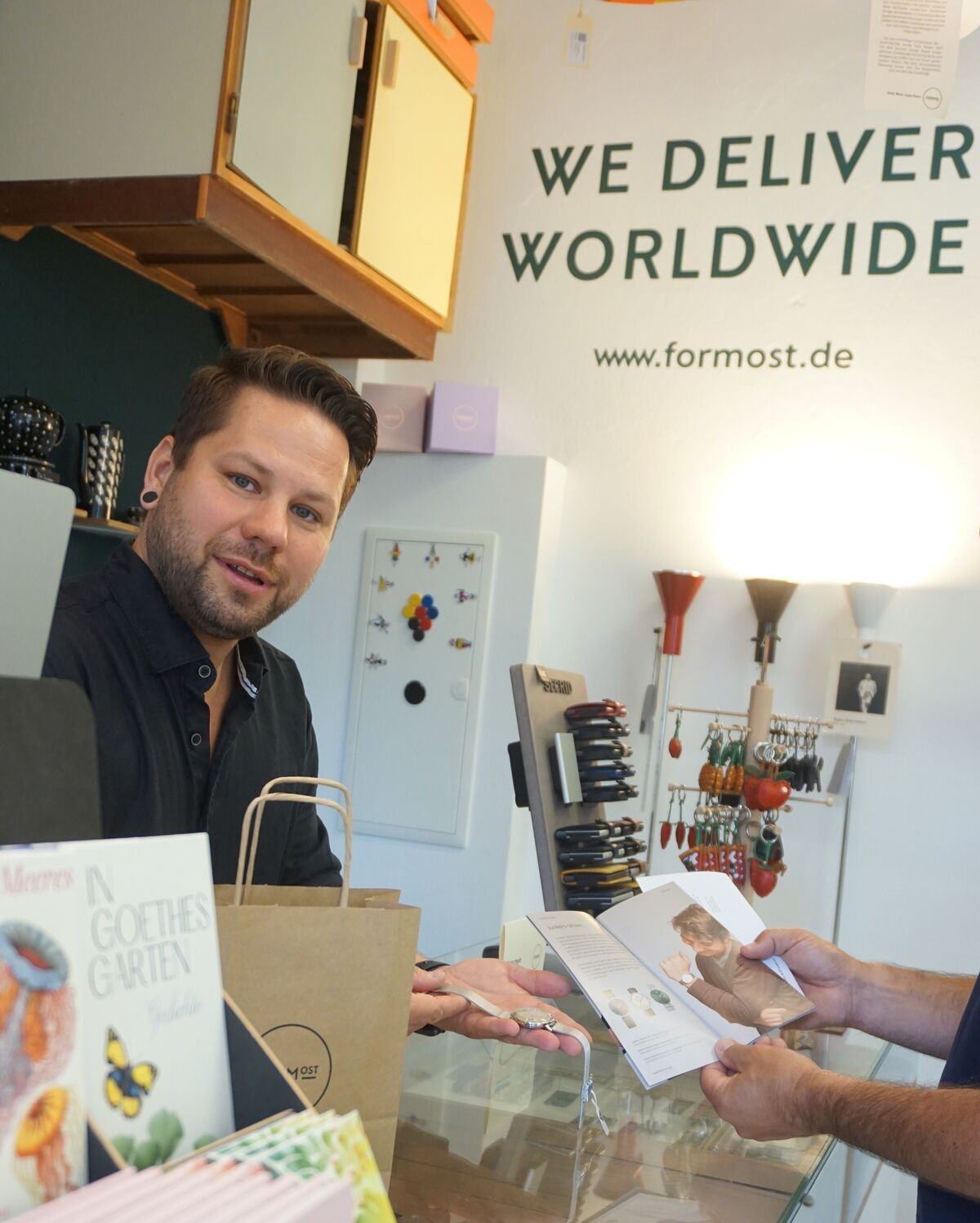 Marcel übergibt einem Kunden eine Uhr und einen Produktkatalog über die Ladentheke, im Hintergrund der Schriftzug „We deliver worldwide"