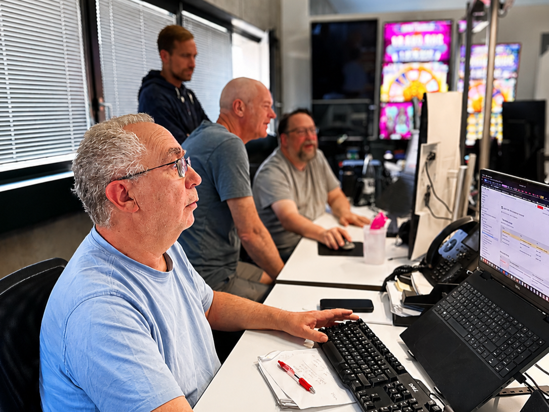 Eingespieltes Team im fachlichen Austausch Vier Männer arbeiten an Schreibtischen mit mehreren Monitoren, im Hintergrund leuchtende Spielautomaten-Displays