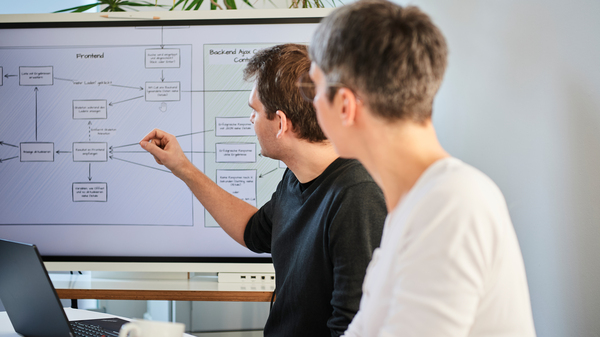 Zwei Personen besprechen technische Abläufe anhand eines Architekturdiagramms auf einem Bildschirm.