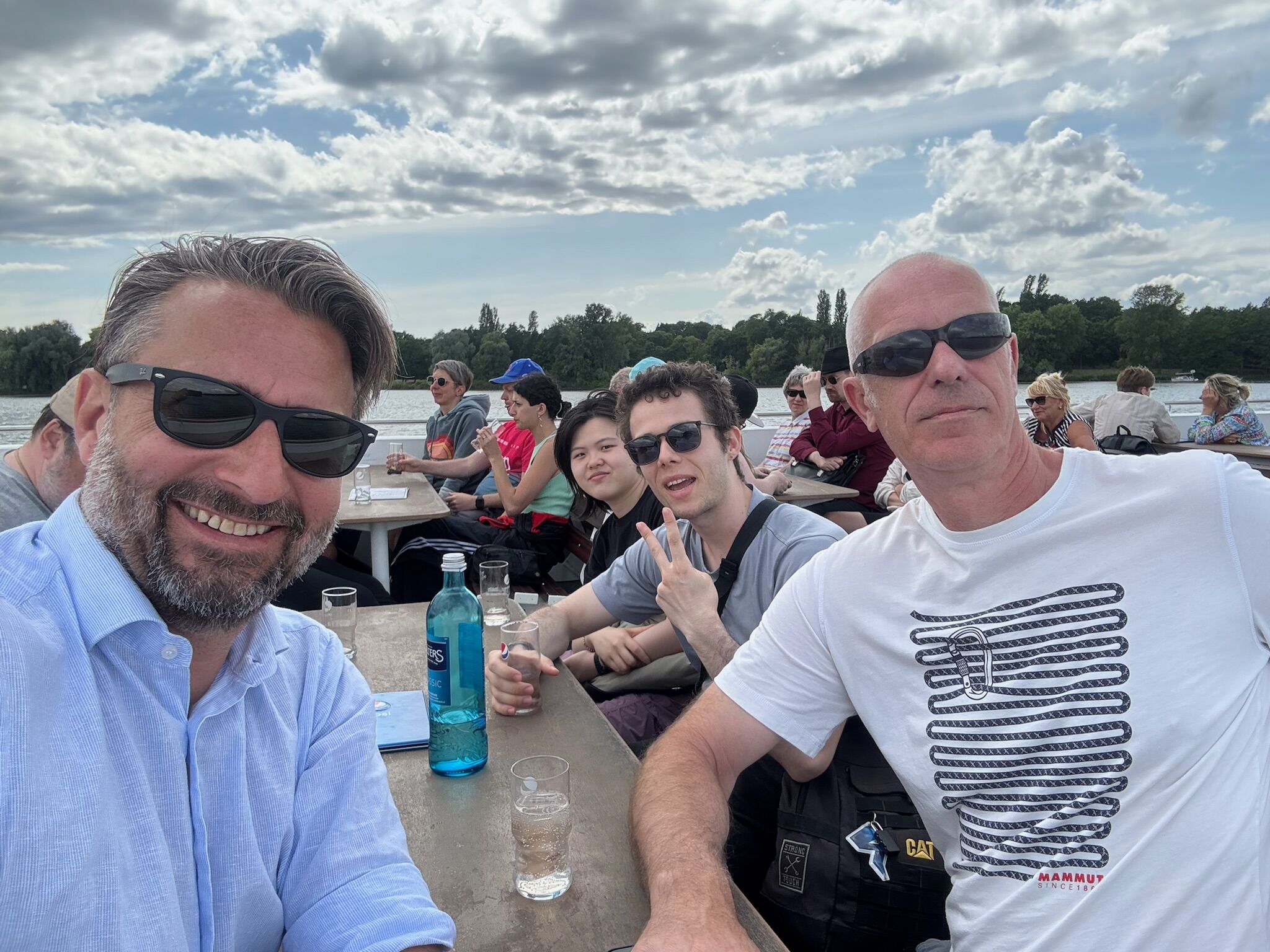 Eine fröhliche Gruppe von Kolleginnen und Kollegen auf einem Ausflugsschiff. Sie tragen Sonnenbrillen, lachen, halten Getränke in der Hand und genießen das sonnige Wetter an Deck.