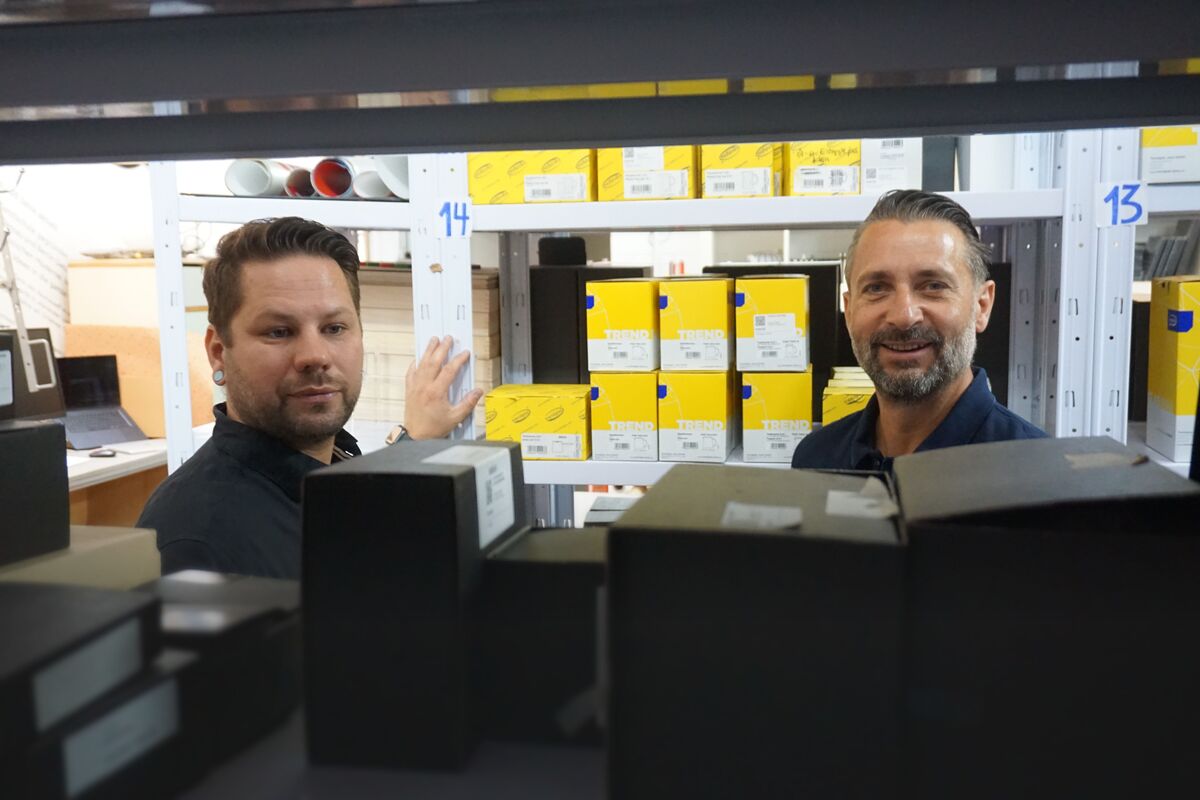 Bestandsprüfung im FORMOST-Lager Marcel und Christian Allihn stehen lächelnd vor einem Lagerregal, im Vordergrund unscharf zwei Archivboxen
