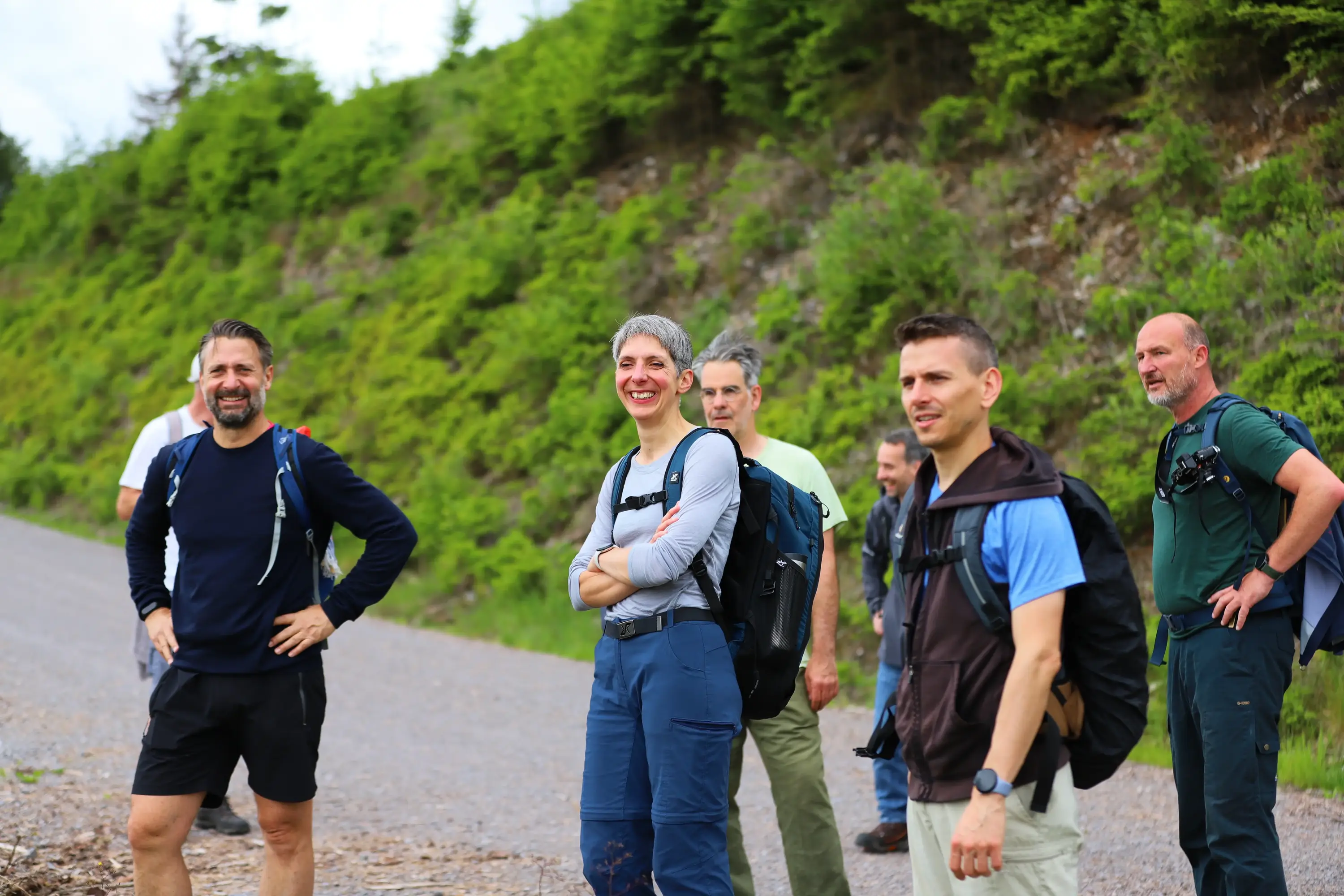 Sechs lachende Personen mit Wanderrucksäcken stehen auf einem Schotterweg vor einem grünen Hang.