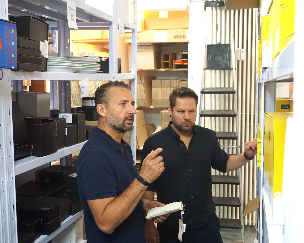 Marcel und Christian Allihn stehen im Lager vor einem Regal und deuten gemeinsam auf ein Fach mit gelben Verpackungsboxen